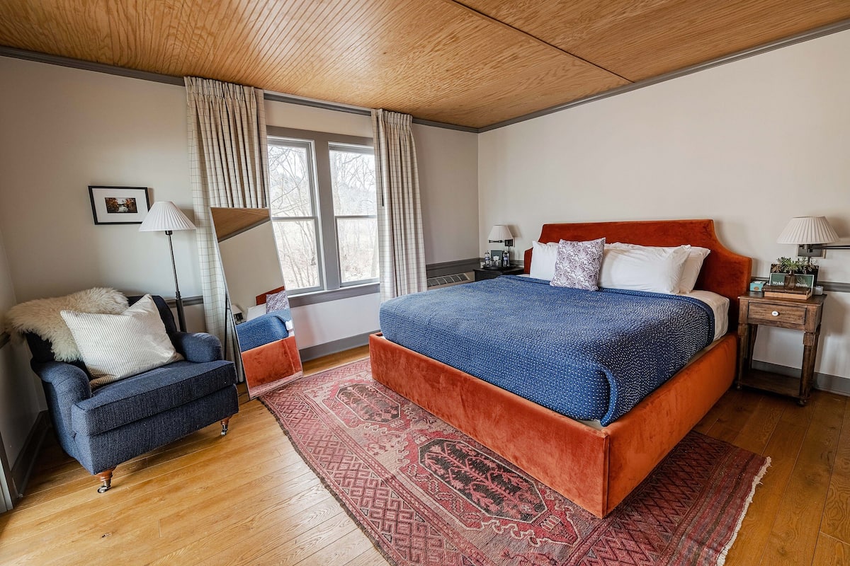 A bedroom with a bed, a chair, a nightstand, a lamp, a window with curtains, and a picture on the wall.