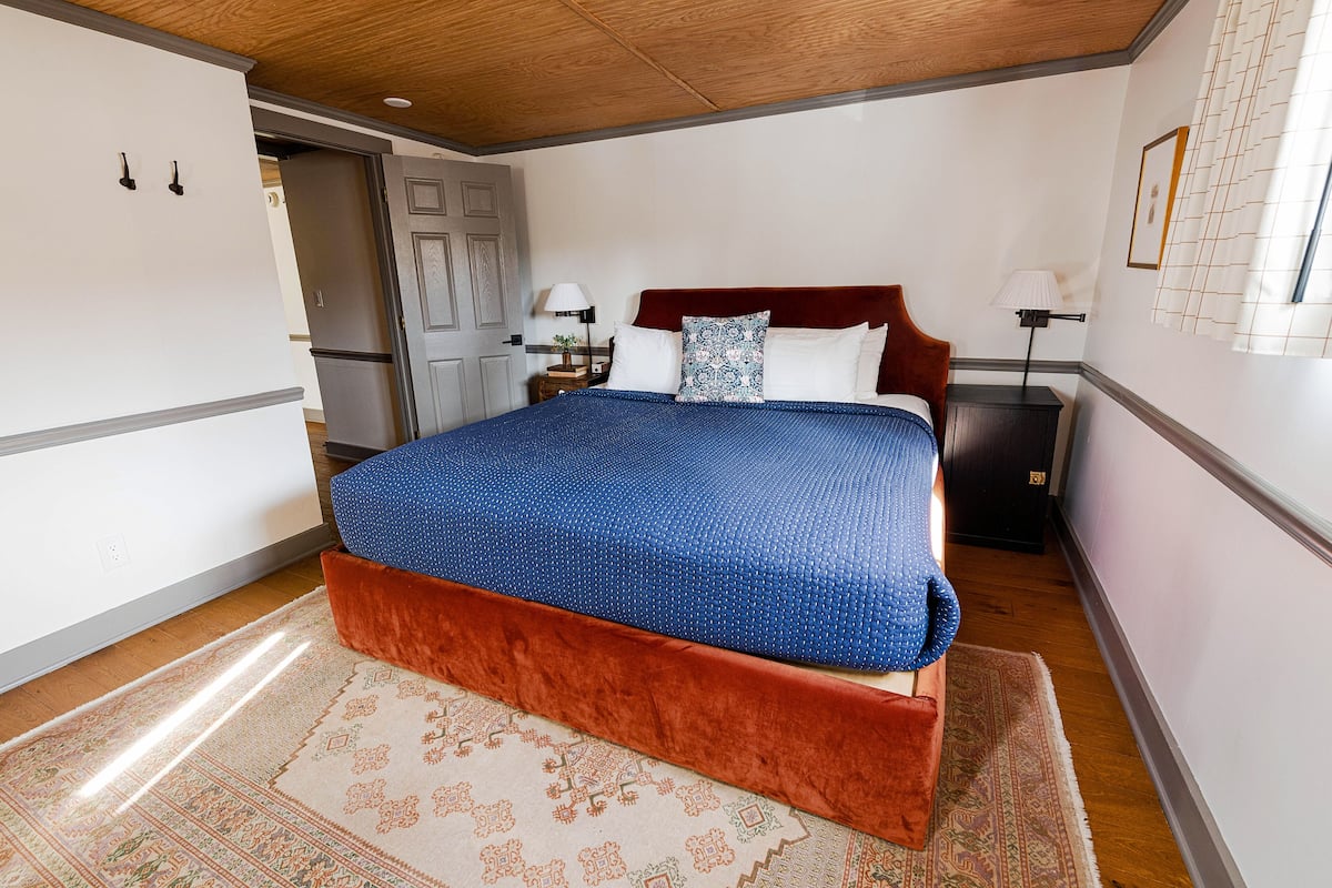 A bedroom with a large bed, a wooden ceiling, a nightstand with a lamp, and a door leading to another room.
