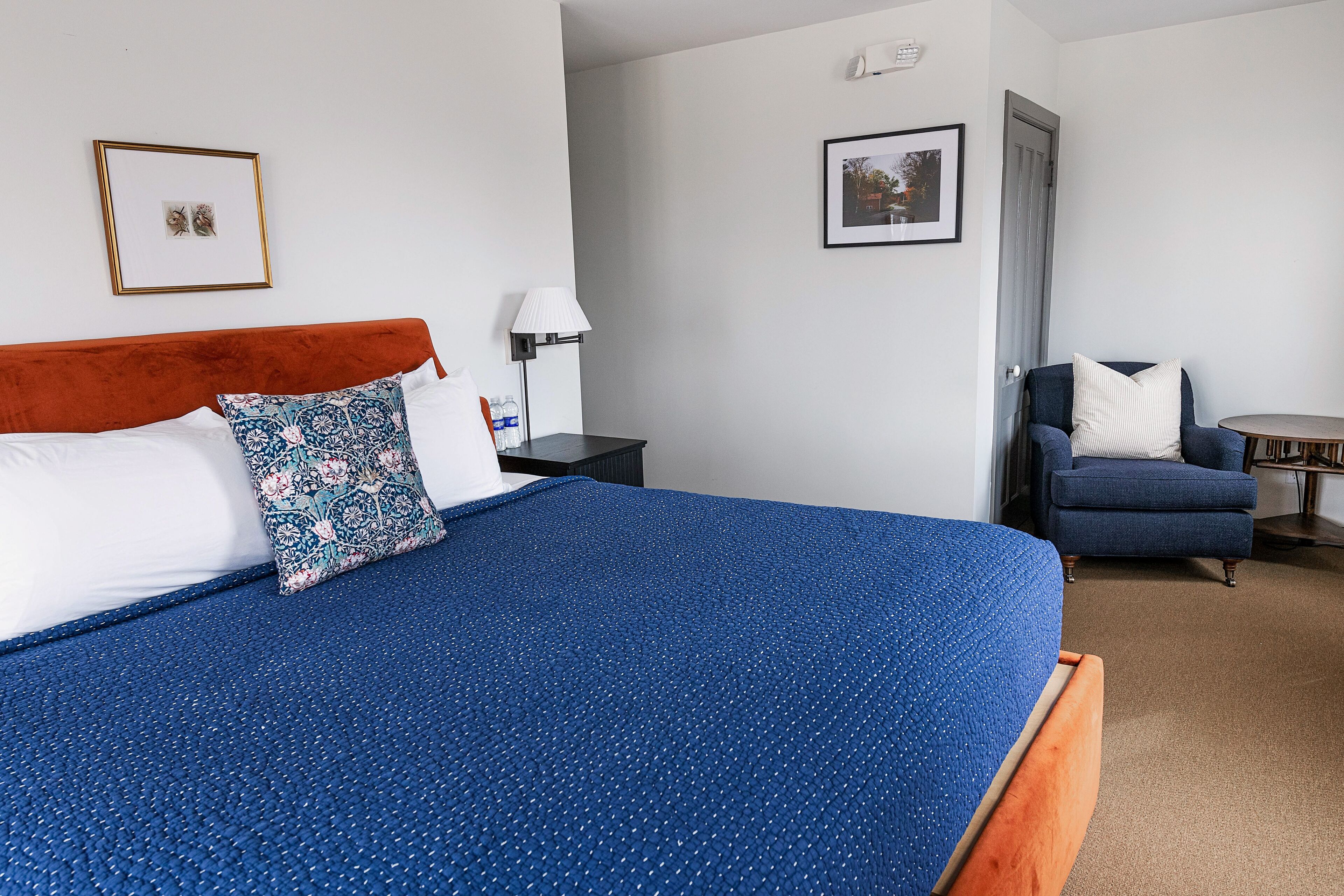 Habitación de hotel ordenada con una cama grande, una silla, una mesita y dos cuadros en las paredes.