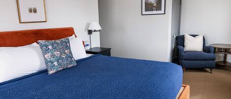 Habitación de hotel ordenada con una cama grande, una silla, una mesita y dos cuadros en las paredes.