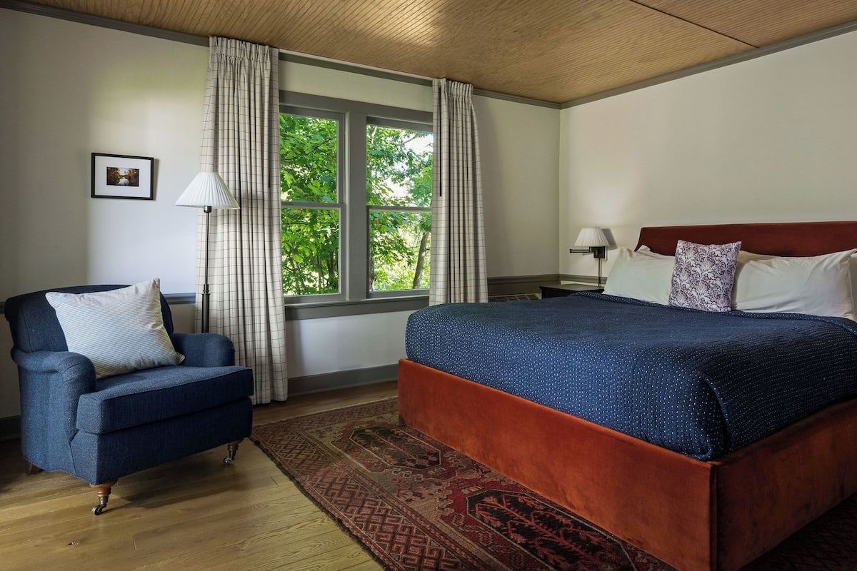 A bedroom with a bed, a chair, a window with curtains, a picture on the wall, and a nightstand with a lamp.