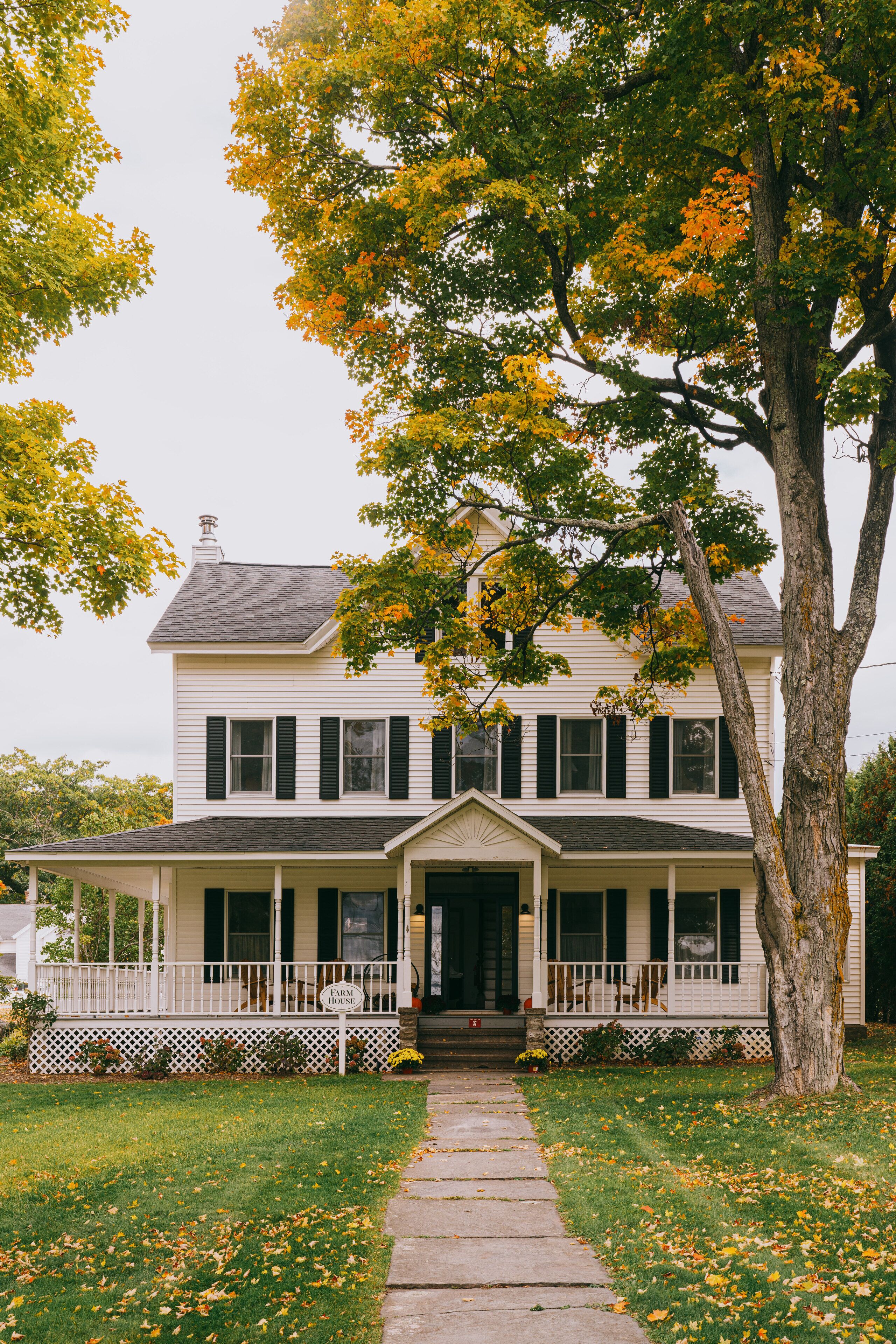 Une maison à deux étages avec un porche à l’avant, une cheminée et un grand arbre devant.