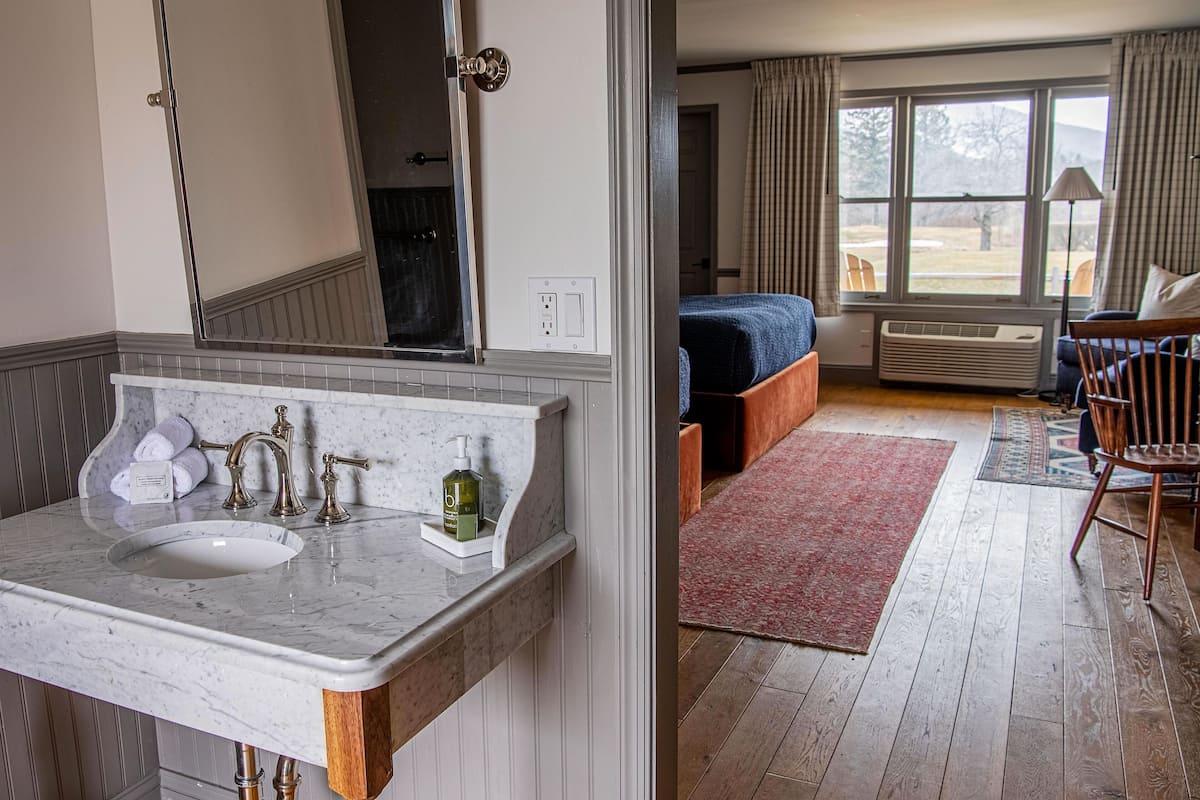 A bathroom with a marble sink, a mirror, and a towel rack. The room has wooden flooring and a view of a bed and a chair through a window.