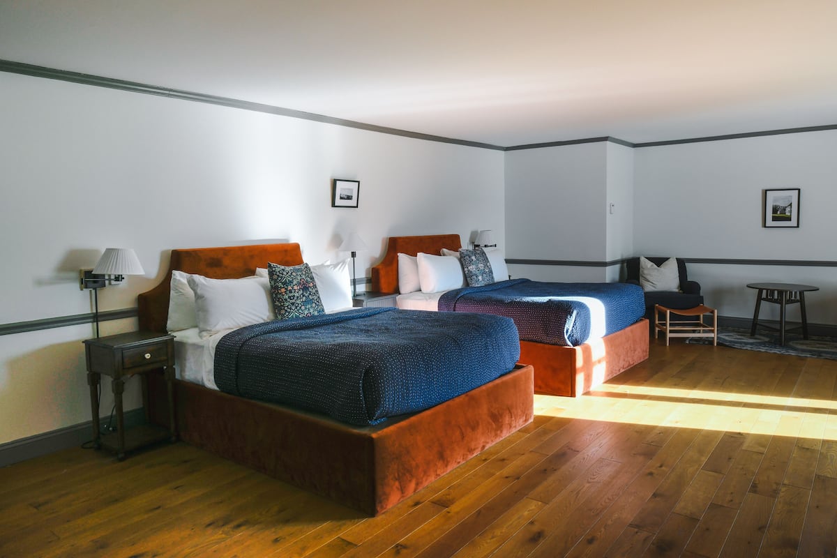 A hotel room with two beds, wooden flooring, and a small table with a lamp.