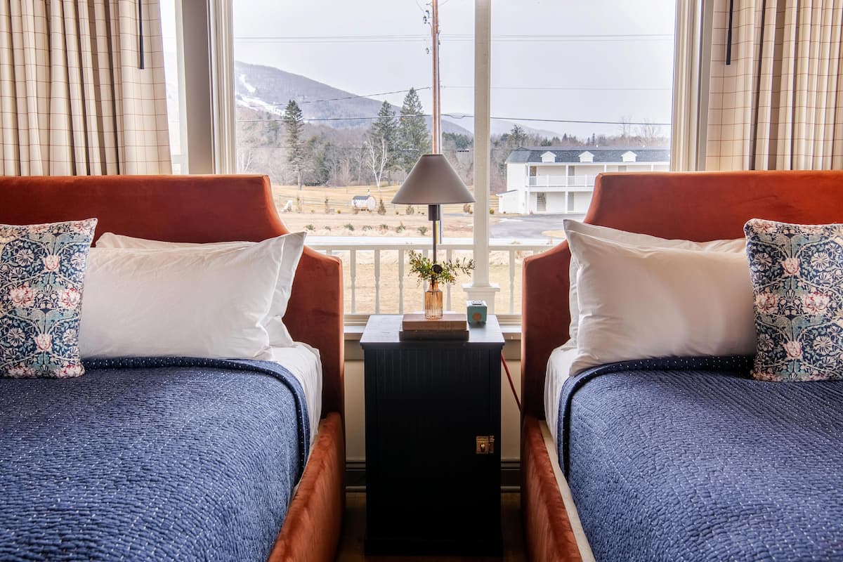 A room with two beds, each with a floral pillow, a small table with a lamp, and a view of a mountainous landscape through large windows.