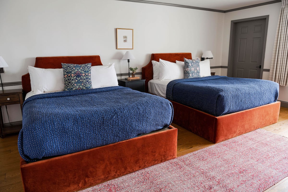 A hotel room with two beds, each with a blue blanket, a red headboard, and white pillows. There is a small table with a lamp and a framed picture on the wall.