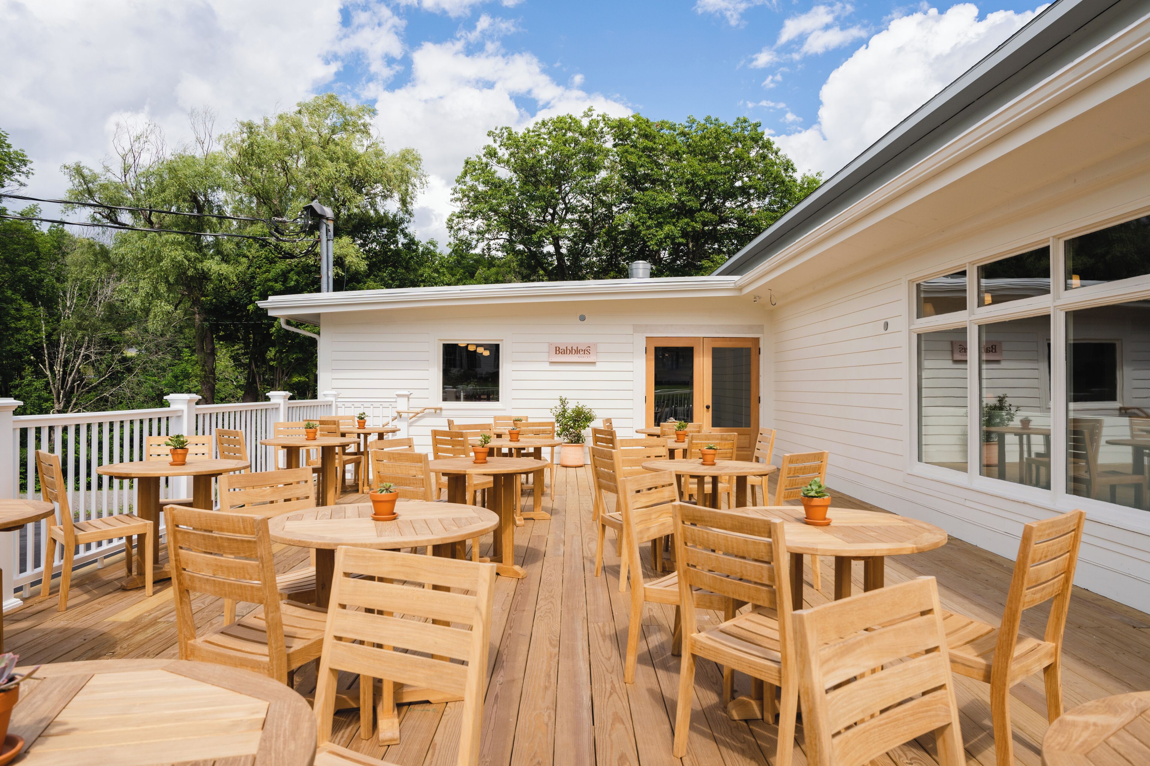 A covered outdoor patio with wooden tables and chairs, a white building with a sign, and trees in the background.