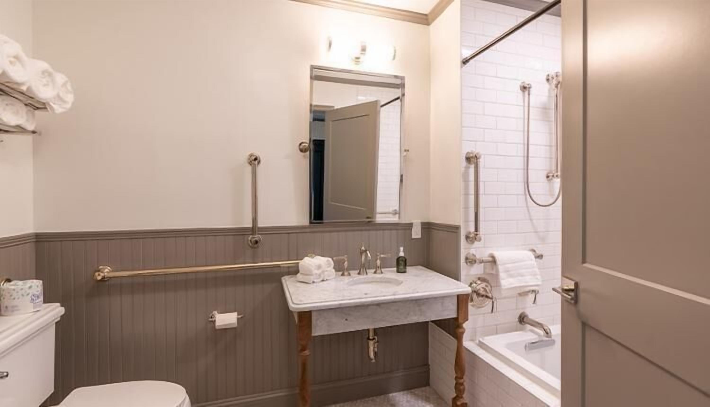 Ein Badezimmer mit Toilette, Waschbecken und Badewanne. Haltegriffe sind in der Nähe der Toilette und des Waschbeckens angebracht.