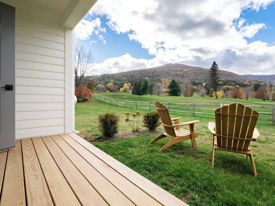 A wooden deck with two Adirondack chairs facing a grassy field and a mountain in the background.