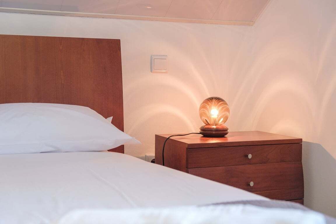 A bedroom with a wooden headboard, a bedside table with a lamp, and a wall-mounted light switch.