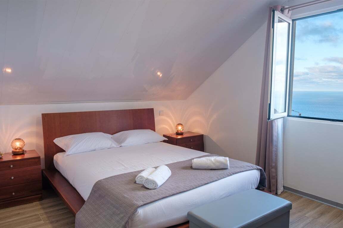 Une chambre à coucher avec un grand lit, des tables de chevet, une commode, un banc et une fenêtre donnant sur la mer.
