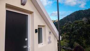 Ein Gebäude mit einer beige farbenen Fassade, einer schwarzen Tür und einem Fenster. An der Wand ist ein Schild mit der Aufschrift 'AL CASA DO GAMBRO' angebracht. Im Hintergrund ist eine gebirgige Landschaft mit viel Grün zu sehen.