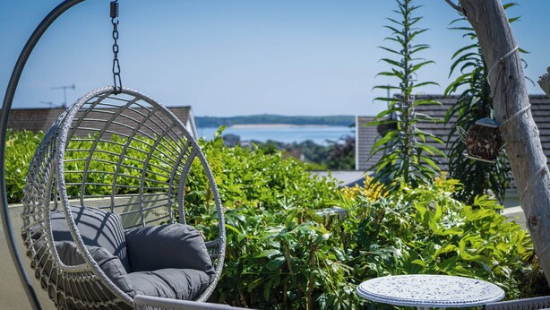A wicker hanging chair with a cushion, a small round table, and lush greenery. - Town Heights - 3 Bedroom Holiday Home - Tenby (Tenby)