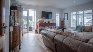 A spacious living room with a large leather sofa, wooden cabinet, and a television mounted on the wall. - Town Heights - 3 Bedroom Holiday Home - Tenby (Tenby)