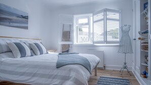 A bedroom with a bed, a window with shutters, a radiator, a patterned rug, and a shelf with items. - Town Heights - 3 Bedroom Holiday Home - Tenby (Tenby)