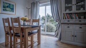 A dining area with a wooden table and chairs, a vase with flowers, and a cabinet with books and dishes. - Town Heights - 3 Bedroom Holiday Home - Tenby (Tenby)