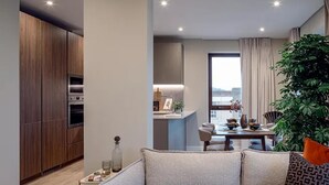 A modern living room with a sofa, a dining area with a table and chairs, a kitchen with wooden cabinets and appliances, and a large window with curtains. - Luxurious 2bed Apartment in Greenford Quays (Greenford)