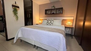 Une chambre d’hôtel bien aménagée, avec un lit, des lampes de chevet, une petite étagère ornée d’objets décoratifs et une porte donnant sur une autre pièce.