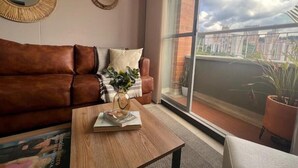 Un salon avec un canapé en cuir marron, une table basse en bois et une grande fenêtre donnant sur un balcon et des immeubles.