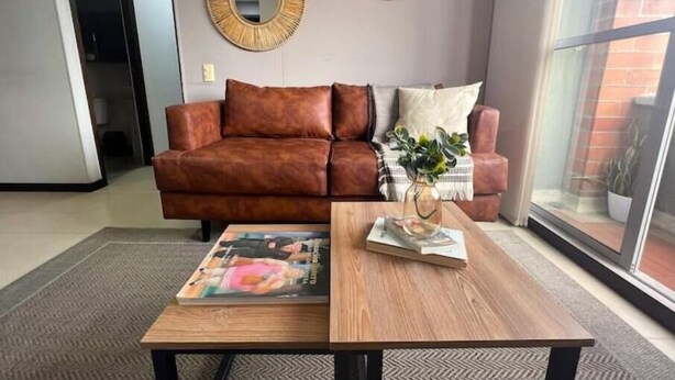 Un salon avec un canapé en cuir marron, deux miroirs muraux, une table basse en bois sur laquelle sont posés des magazines, et une vue dégagée sur l’extérieur grâce à une grande fenêtre.