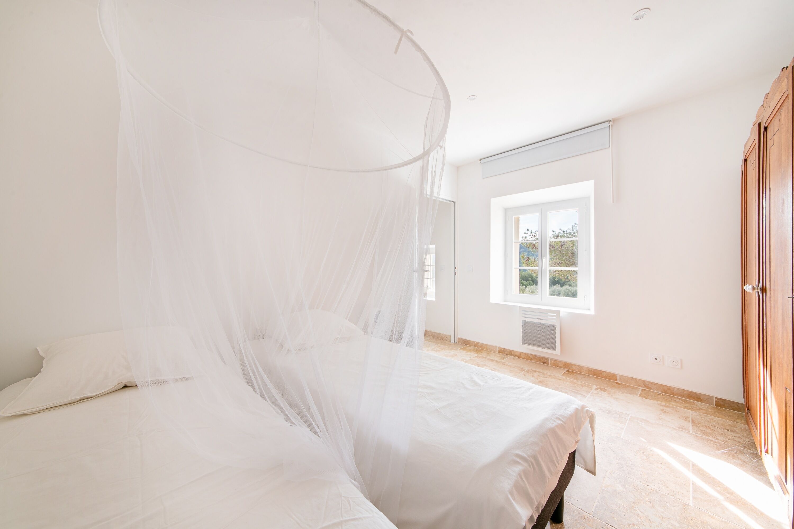 A bedroom with a bed under a mosquito net, a window with a view, and a wooden wardrobe.