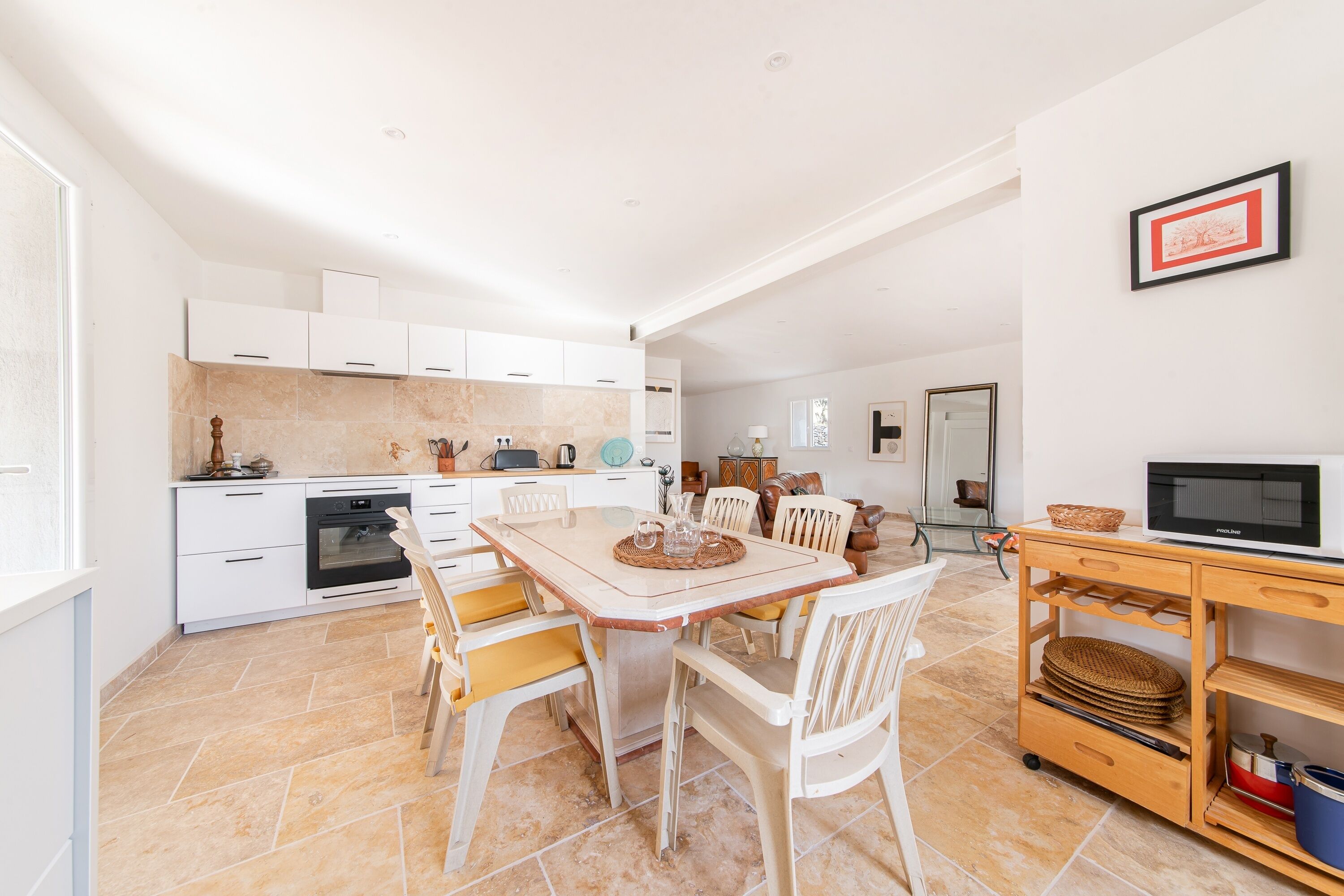 A modern kitchen with white cabinetry, a dining table with chairs, a wooden cart with kitchen items, and a microwave on a wooden shelf.