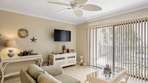 A living room with a beige sofa, a glass coffee table, a white console table, a ceiling fan, a TV mounted on the wall, and a view through a window with blinds. - Beachside Charm! Just A Short Stroll To Miles Of Pristine Beach. (St. Augustine)