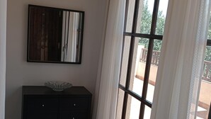 Ein Zimmer mit einem dunklen Holzschrank, einer Glasbowl darauf und einem großen Fenster mit weißen Vorhängen.