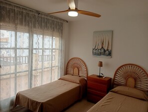 Een slaapkamer met twee eenpersoonsbedden, een plafondventilator, een nachtkastje met een lamp en een schilderij van zeilboten aan de muur.