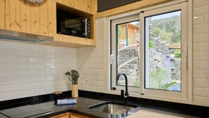 A modern kitchen with wooden cabinets, a black countertop, a stainless steel sink, and a window with a view of a stone building and greenery. - Casas Maravilha - Piscina by Homie (Calheta)
