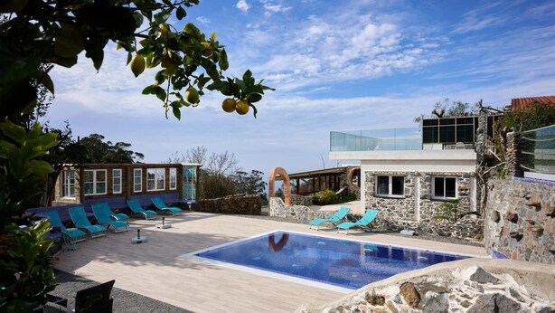 A pool area with a rectangular pool, stone walls, and blue lounge chairs. - Casas Maravilha - Piscina by Homie (Calheta)