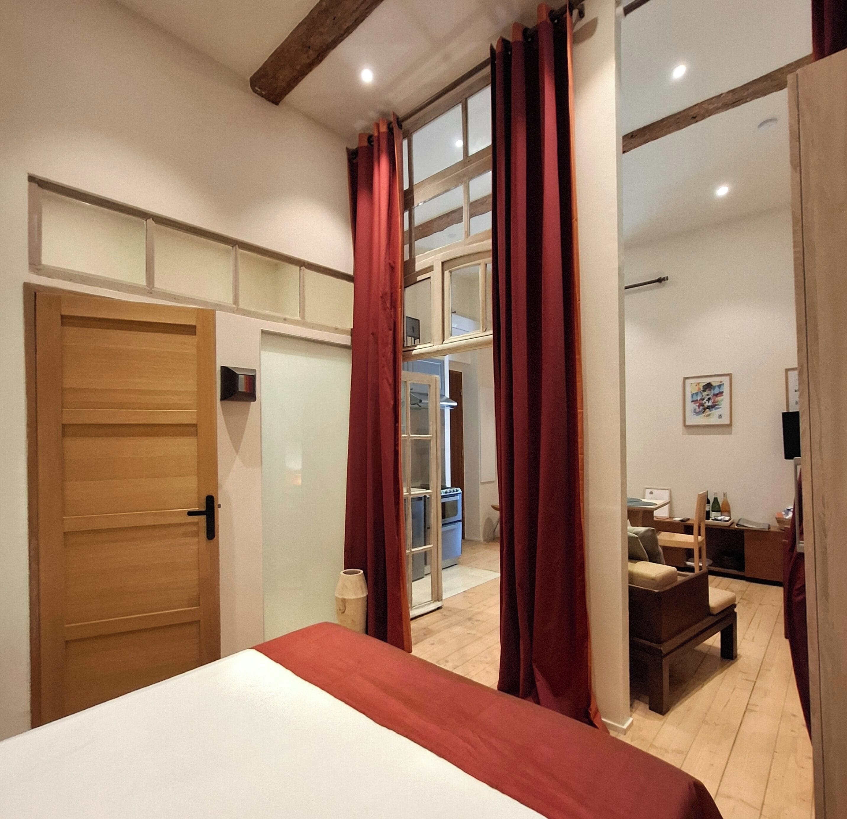 Une chambre avec une porte en bois, un lit recouvert d’une couverture rouge, une grande fenêtre avec des rideaux rouges et une vue sur la cuisine.
