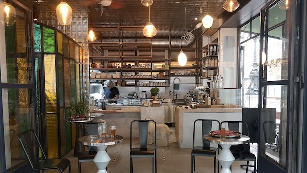 Un café con mesas redondas, sillas y una barra donde una persona prepara comida.
