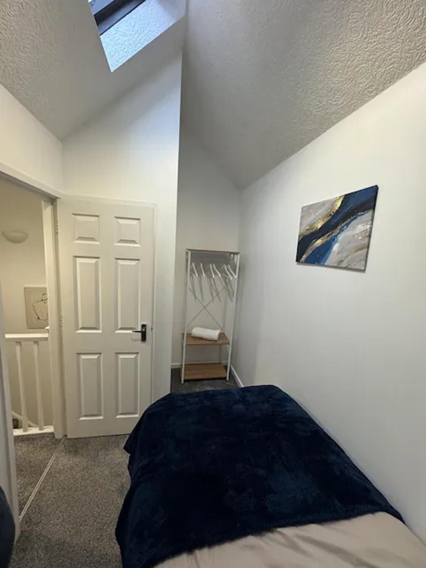 A narrow room with a bed, a white door, a coat rack, and a painting on the wall.