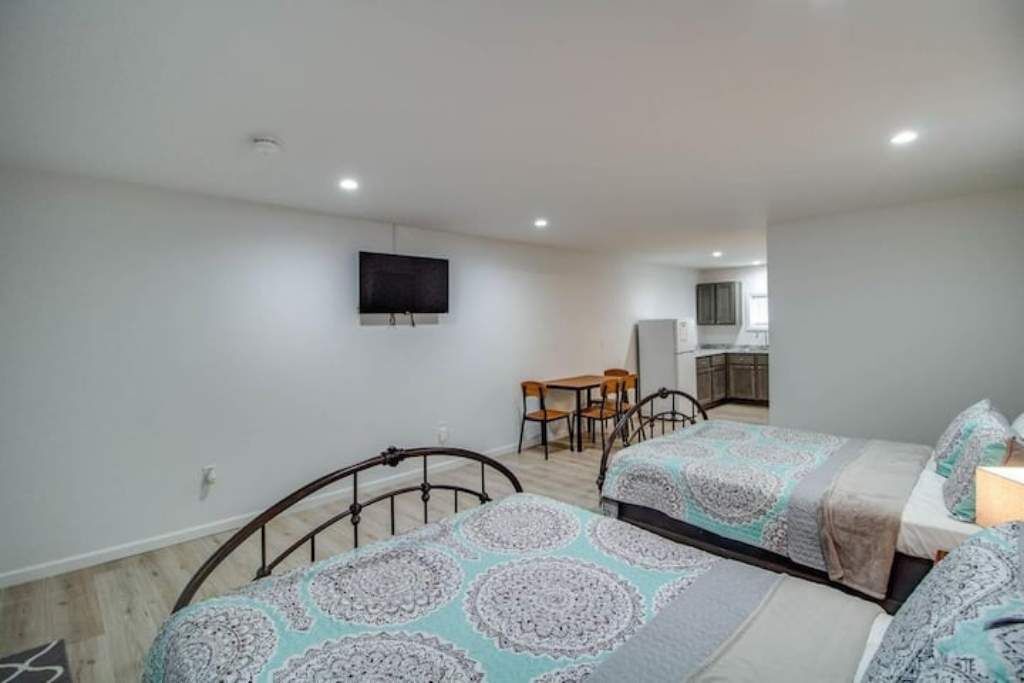A bedroom with two beds, a TV mounted on the wall, and a small kitchen area in the background.