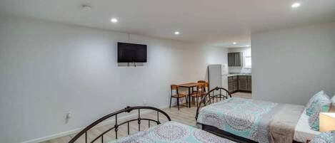 A bedroom with two beds, a TV mounted on the wall, and a small kitchen area in the background.