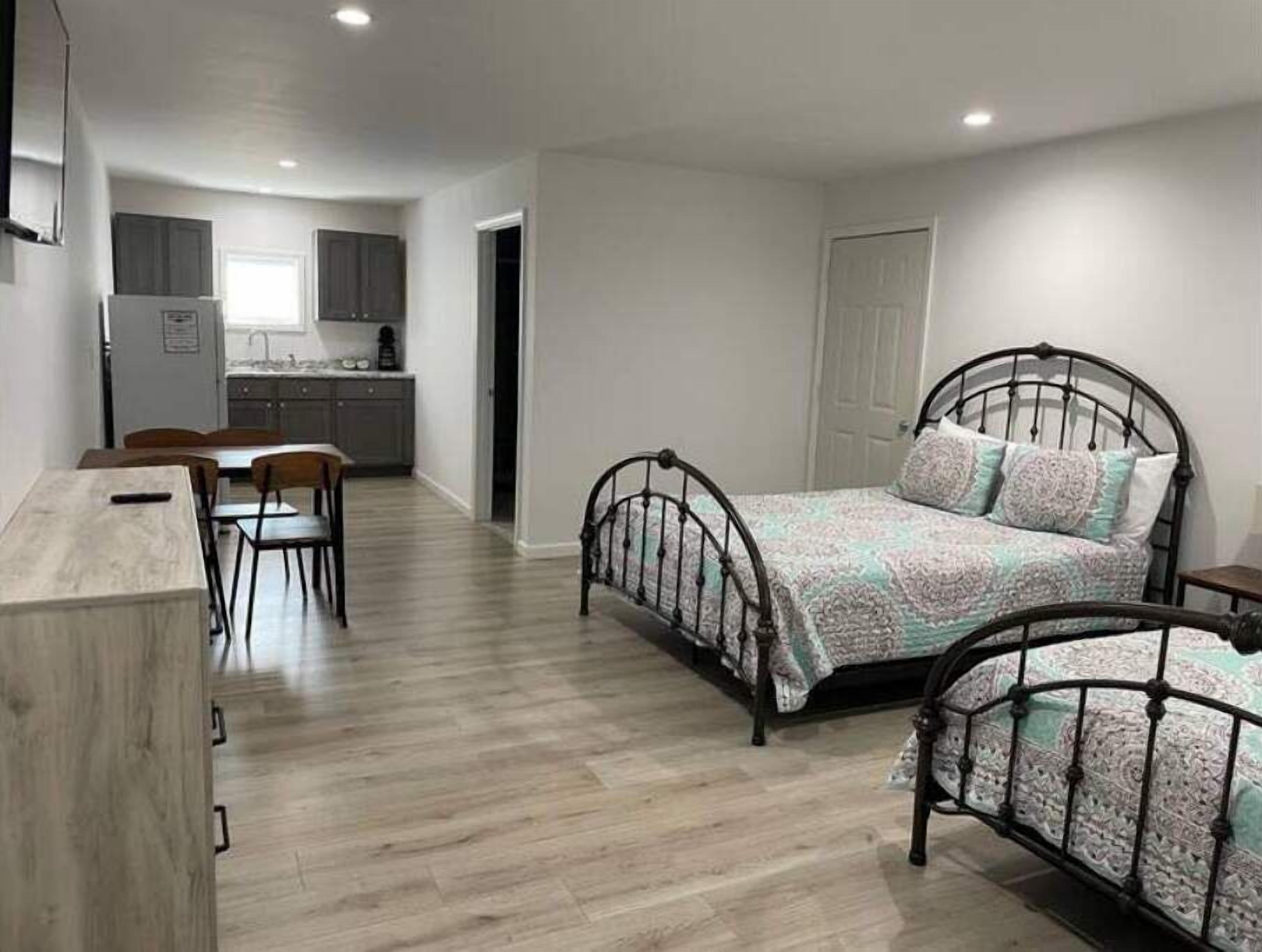 A bedroom with a bed, a desk, and a chair. There is a kitchenette in the background.