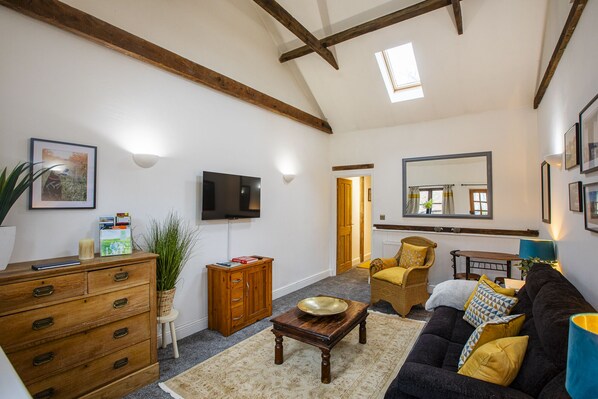 Ein geräumiges Wohnzimmer mit hoher Decke, Holzbalken, einem Flachbildfernseher, einem großen Spiegel und einem gemütlichen Sofa.