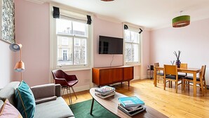 Una sala moderna con un sofá, una mesa de centro con libros, un televisor, una zona de comedor con sillas y una mesa, y ventanas amplias con persianas.