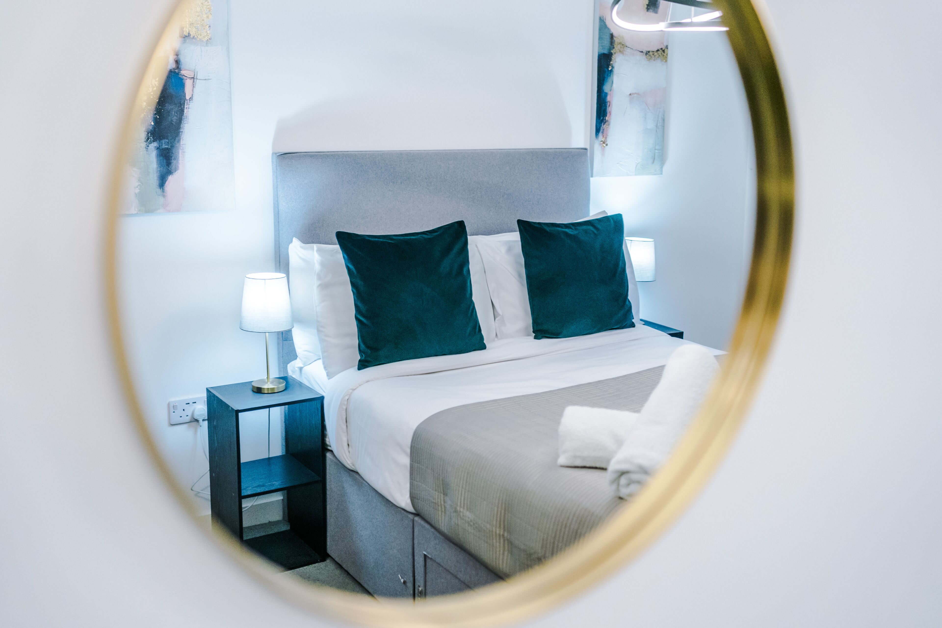 Un lit bien fait, avec une literie blanche et turquoise, une lampe de chevet et une petite table de nuit en bois.
