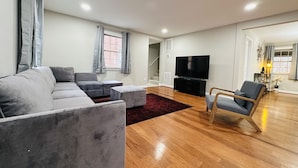A modern living room with a grey sectional sofa, a wooden floor, a flat-screen TV on a black stand, a wooden chair, and a staircase leading to another level.
