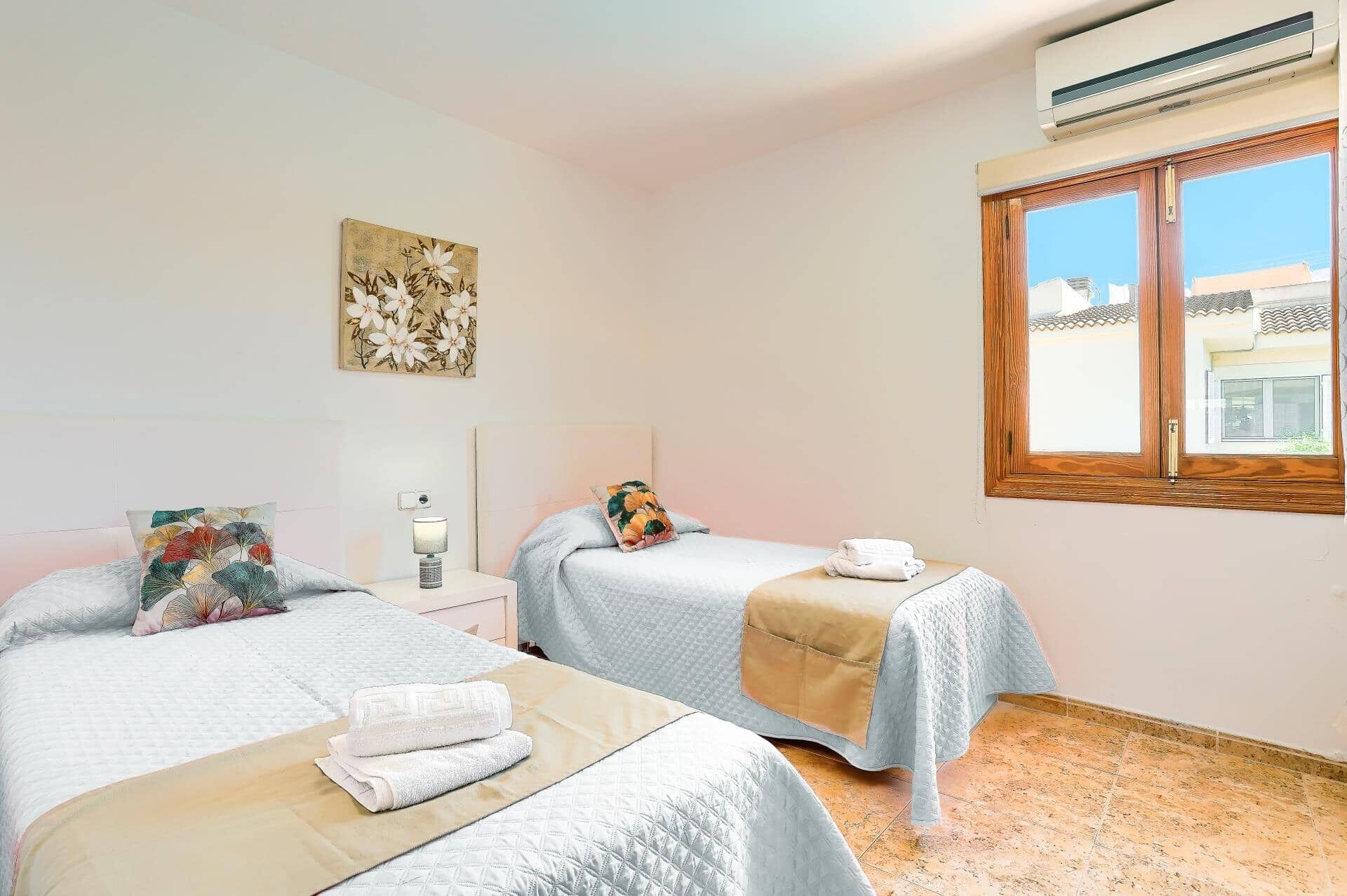 A hotel room with two beds, a nightstand with a lamp, a wall-mounted air conditioner, and a window with a view of buildings.