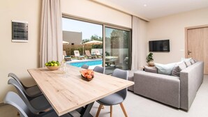 Un salon moderne avec vue sur la piscine, une table à manger avec des fruits et du vin, et un canapé gris.