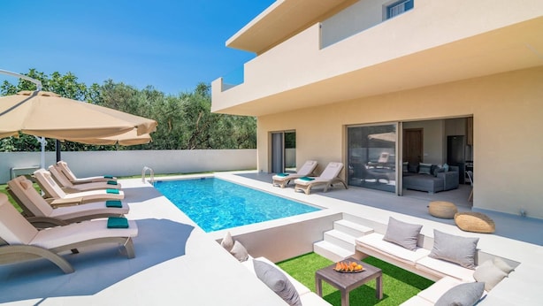 Un espace piscine extérieur moderne, doté de chaises longues et d’un canapé, d’une petite table avec des fruits et d’un salon fermé par des baies vitrées.