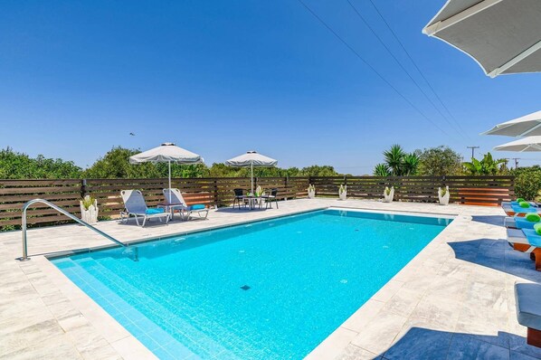 A rectangular swimming pool with a tiled deck, surrounded by lounge chairs and umbrellas.