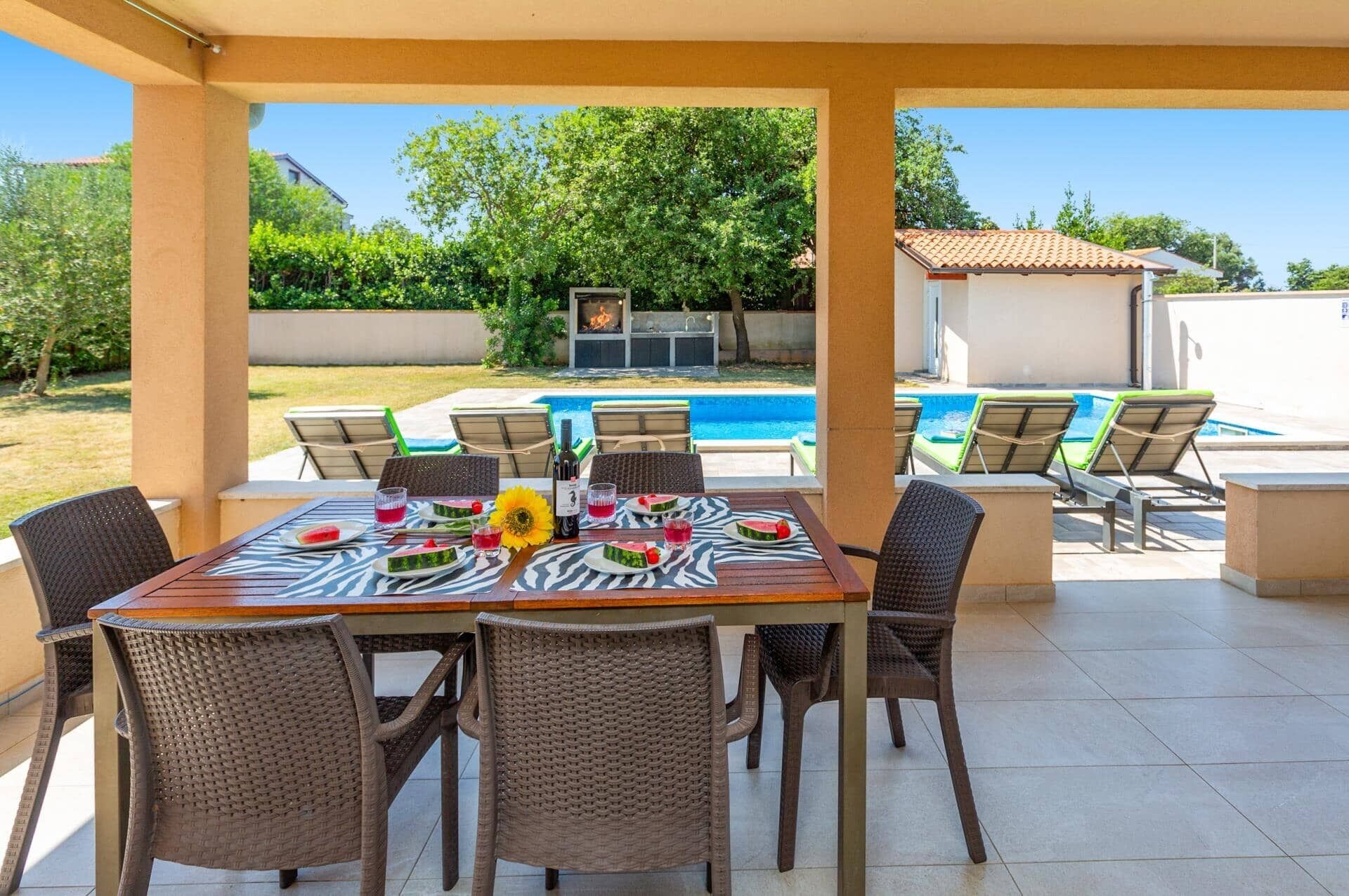 An outdoor patio with a table set for dining, surrounded by wicker chairs, overlooking a swimming pool and a backyard.