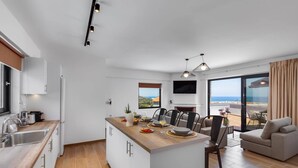 A modern kitchen with a central island, a dining area with chairs, a sofa, and a large window with a view. - Villa Annalena by Villa Plus (Malia)