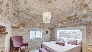 Un dormitorio con pared de piedra, una cama con ropa de cama rosa, una silla, un banco, una lámpara colgante y una ventana con contraventanas.