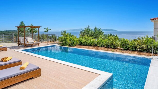 Une piscine bordée de chaises longues et surmontée d’une pergola, d’où l’on peut admirer une mer d’un bleu limpide et une terre lointaine.