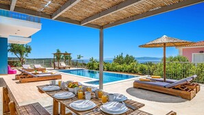 Un espace piscine extérieur avec des chaises longues en bois, une table à manger dressée avec de l’argenterie et des boissons, et une vue sur la mer.